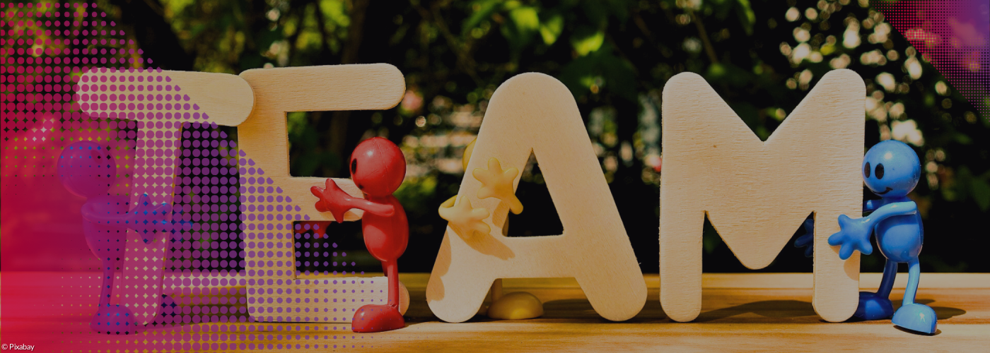 Holzbuchstaben TEAM vor Blätterhintergrund und kleine Figuren, die sich an den einzelnen Buchstaben festhalten.